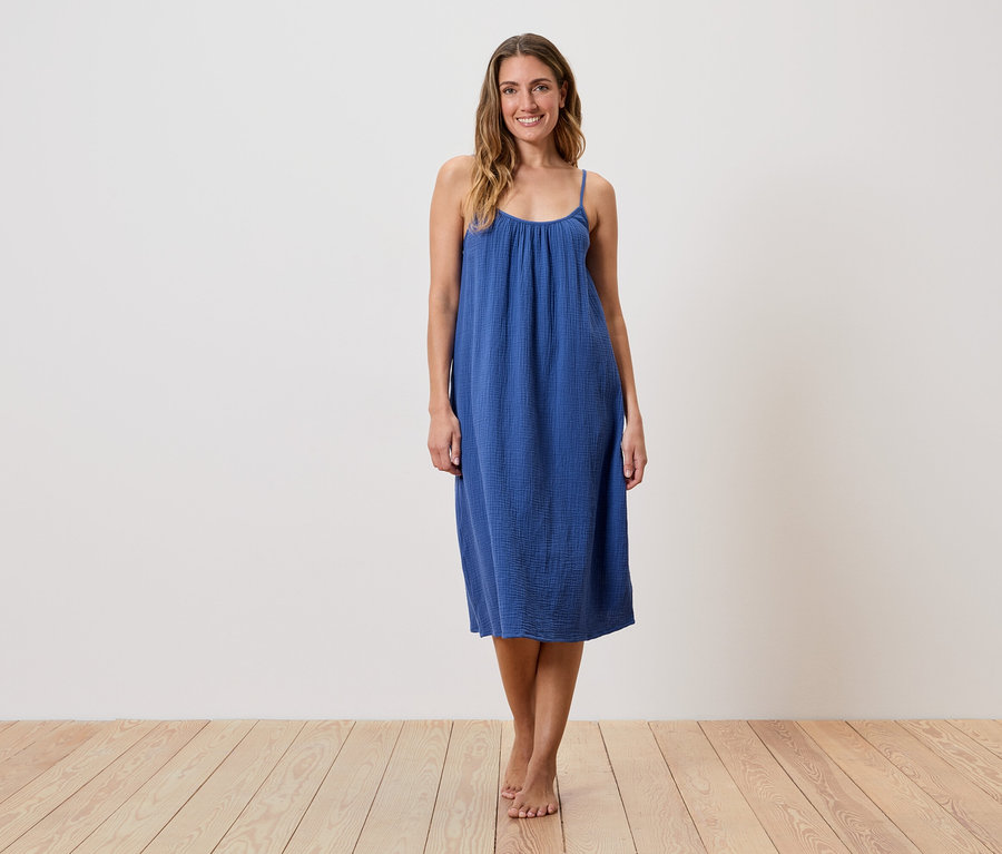 Une femme se tient pieds nus sur un plancher en bois et porte une robe bleue sans manches.
