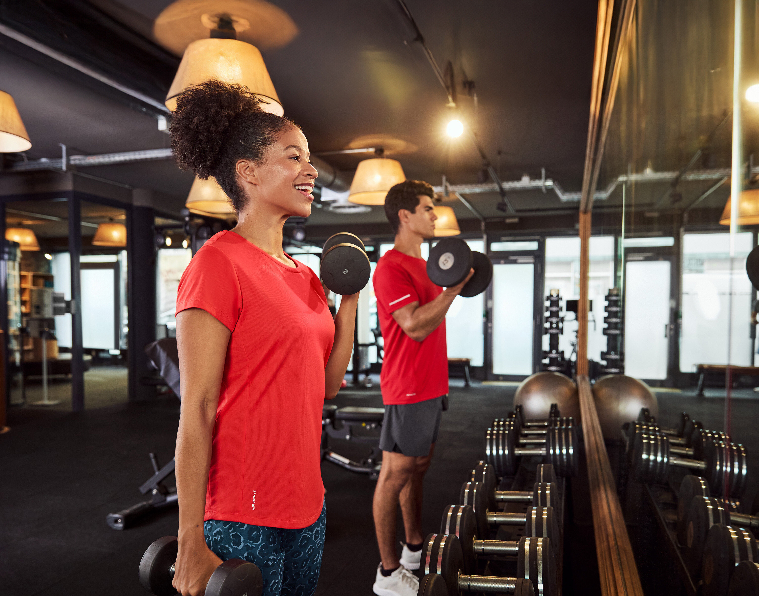 Une femme et un homme font des exercices d'haltères dans une salle de sport devant un miroir.