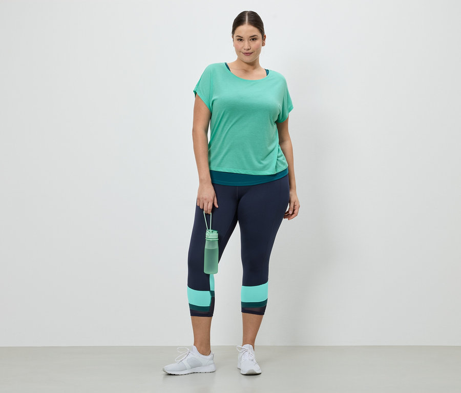 Une femme portant des vêtements de sport composés d'un t-shirt vert, d'un legging bleu marine et de baskets blanches tient une bouteille d'eau verte.