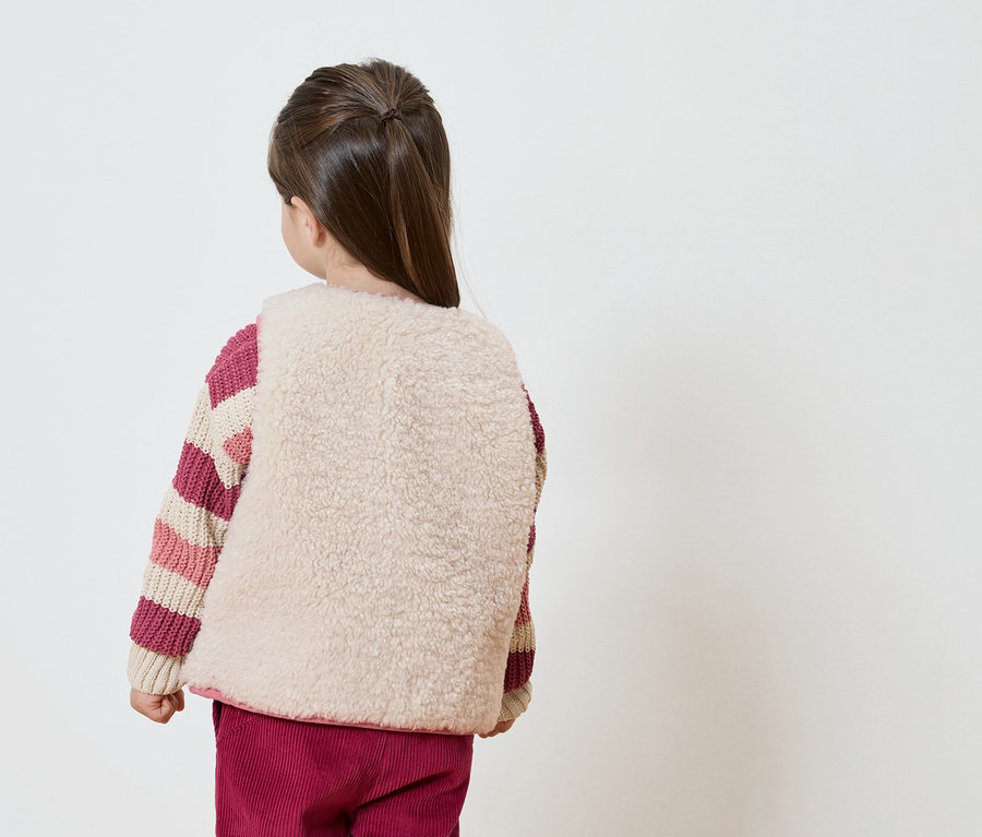 Vue de dos d'une fillette avec une queue de cheval, portant un gilet beige sur un pull rayé et un pantalon bordeaux.