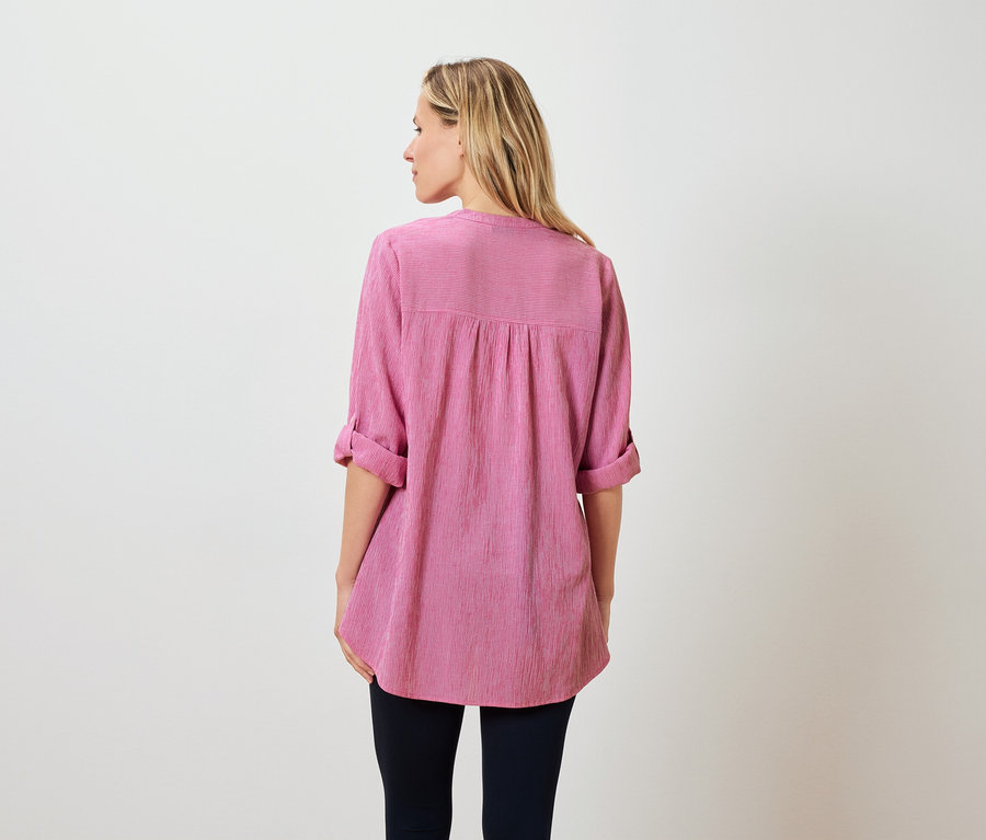 Vue de dos d'une femme aux cheveux blonds mi-longs, vêtue d'un chemisier rose et d'un pantalon foncé.