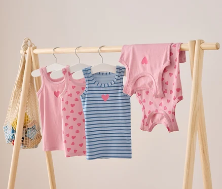 Des débardeurs pour enfants roses et bleus rayés avec des motifs de cœur sont suspendus à une barre en bois.