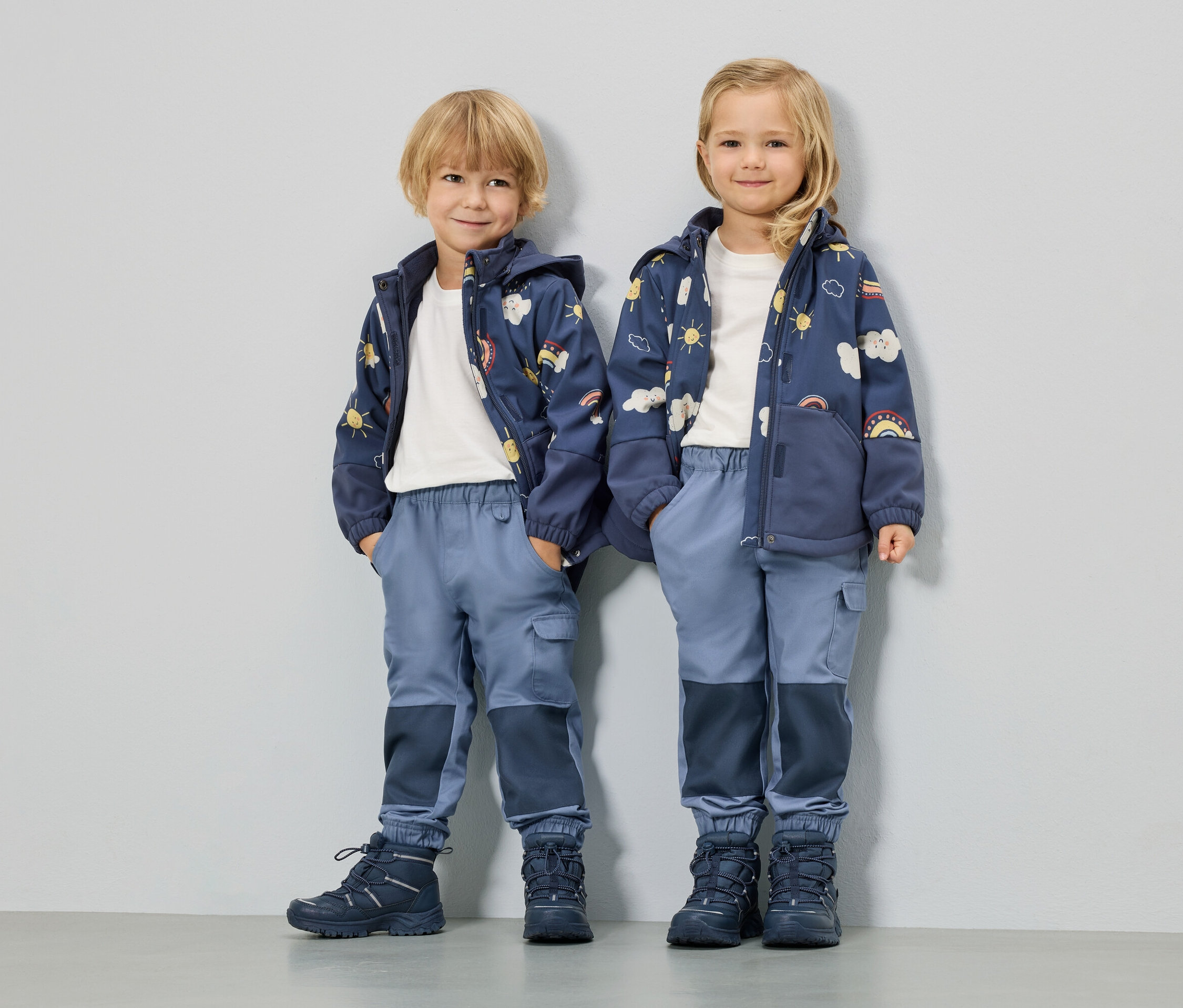 Deux enfants se tiennent côte à côte, portant des vestes bleues avec des motifs de soleil, de nuages et d'arc-en-ciel, des pantalons bleus et des chaussures assorties.