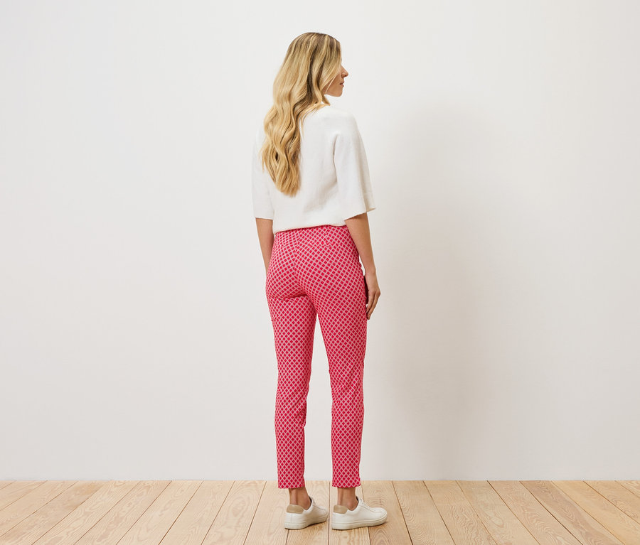 Vue arrière d'une femme aux cheveux blonds ondulés portant un chemisier blanc et un pantalon à motifs rose et rouge avec des chaussures blanches.