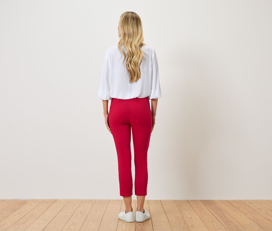 Vue arrière d'une femme aux cheveux blonds ondulés portant un chemisier blanc et un pantalon rouge.