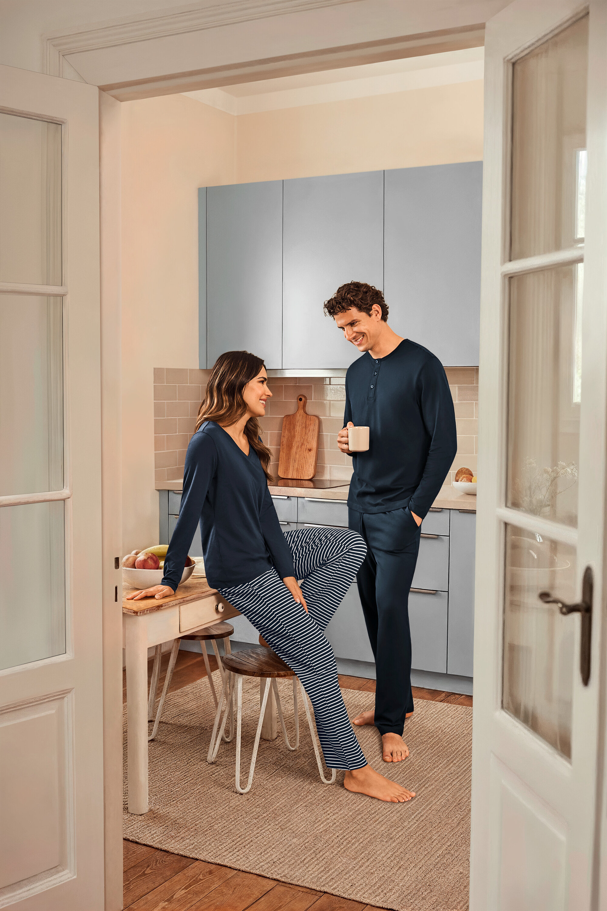 Dans une cuisine, une femme assise sur une table portant un pyjama bleu marine rayé et un haut bleu marine et un homme tenant une tasse et portant un pyjama bleu marine.