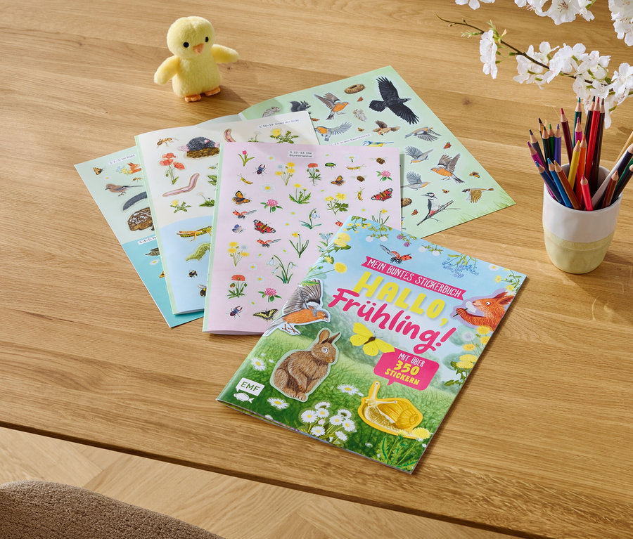 Sur une table en bois se trouvent un poussin jaune, des livres d'autocollants et des crayons de couleur dans une tasse.