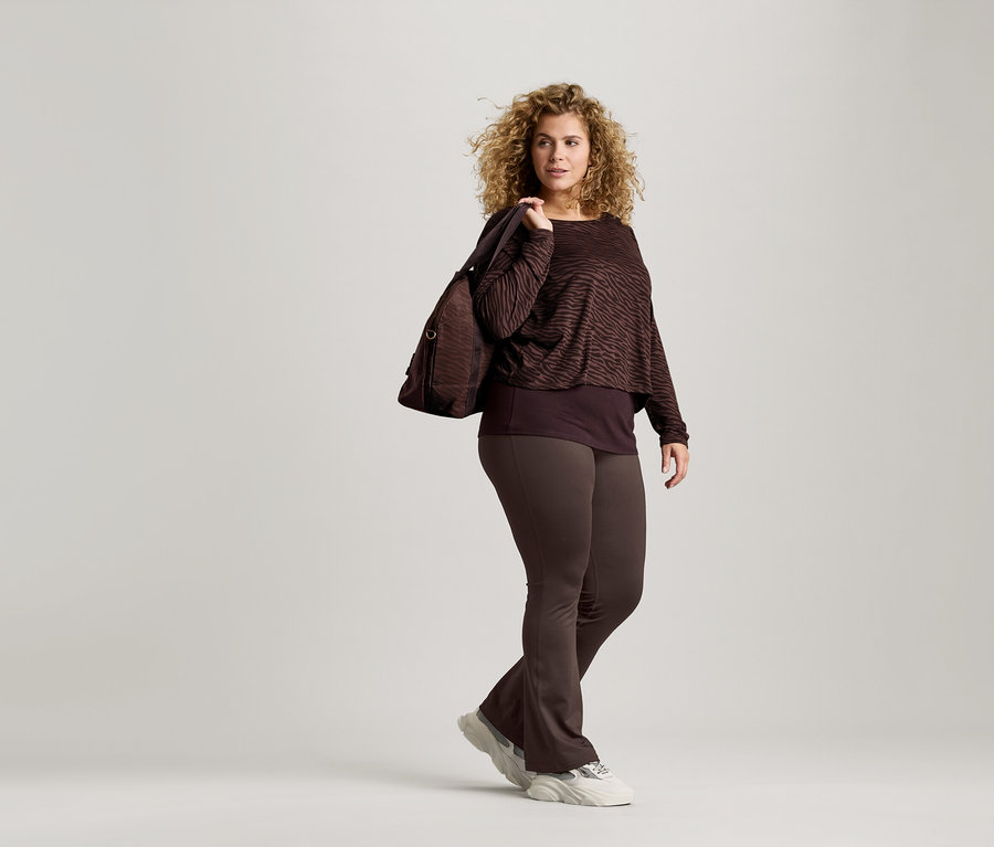 Une femme aux cheveux bouclés porte un haut imprimé zébré marron, un legging marron et des baskets. Elle porte un sac marron sur l'épaule.