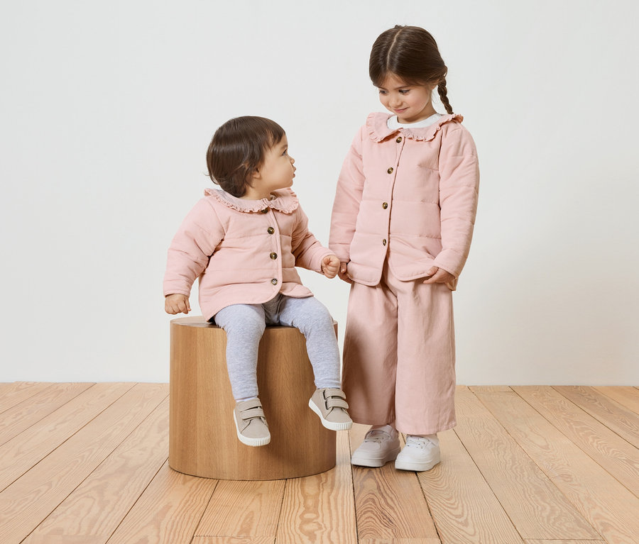 Deux fillettes, l'une assise sur un tabouret en bois, toutes deux vêtues de vestes roses à col à volants.
