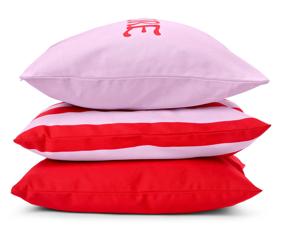 Trois coussins sont empilés : un coussin rose avec une inscription, un coussin rayé rouge et blanc et un coussin rouge.