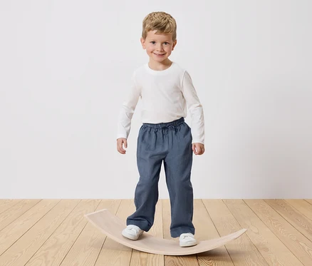 Un garçon souriant aux cheveux blonds se tient debout sur une planche d'équilibre en bois. Il porte un t-shirt blanc à manches longues et un pantalon bleu.