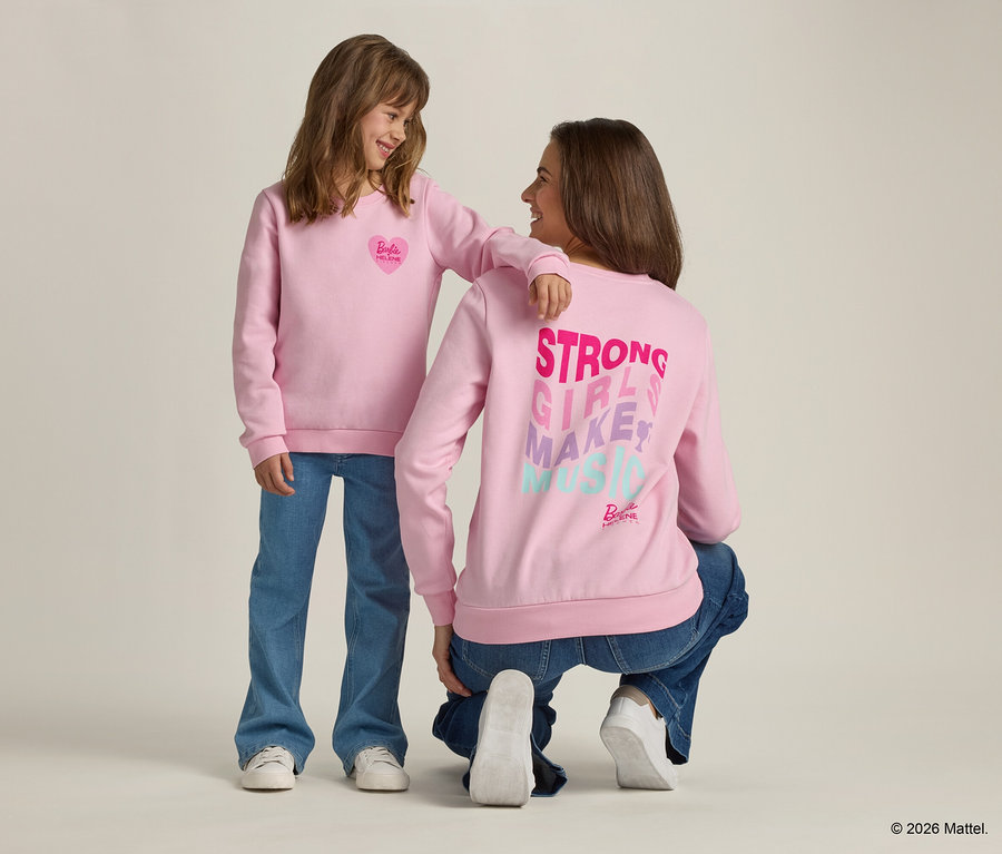 Une fille avec le bras sur l'épaule d'une femme accroupie, toutes deux vêtues de pulls roses et de jeans.