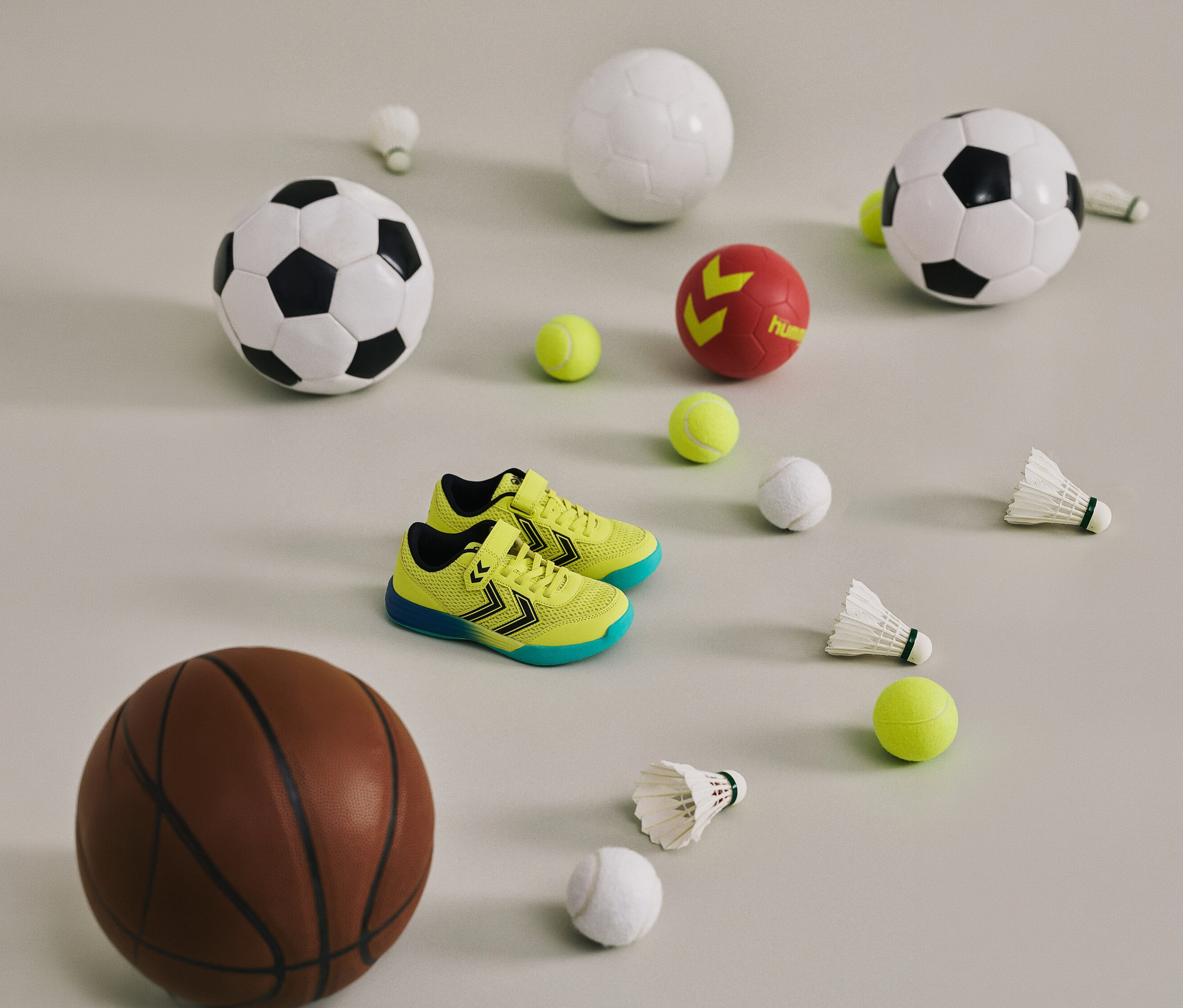 Chaussures jaunes Multiplay Flex VC JR hummel® entre des ballons et des volants sur un fond clair.