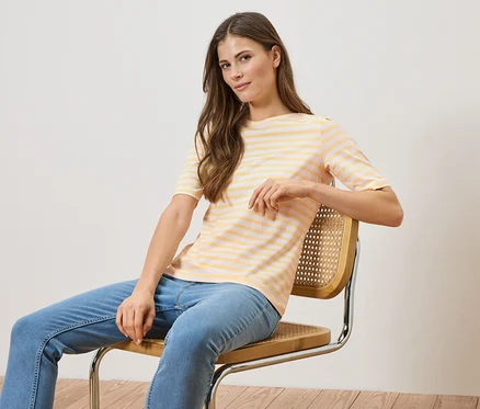 Une femme est assise sur une chaise, portant un t-shirt rayé jaune et blanc et un jean bleu.