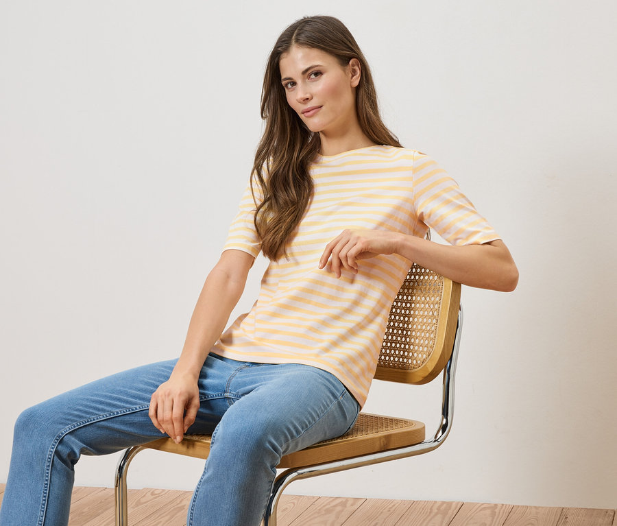 Une femme est assise sur une chaise, portant un t-shirt rayé jaune et blanc et un jean bleu.