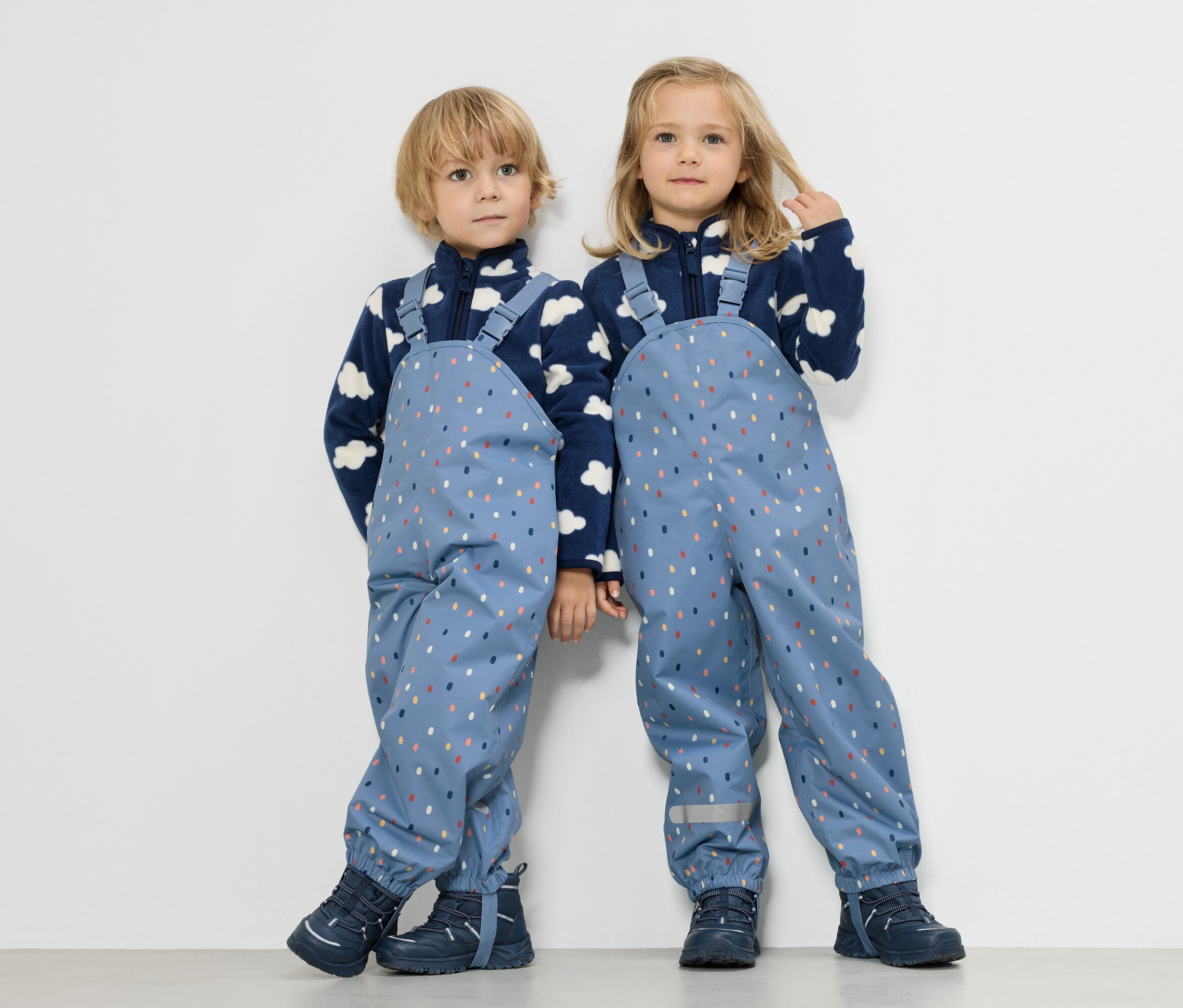 Deux enfants se tiennent côte à côte, un garçon et une fille, tous deux vêtus de vestes polaires bleues à motifs de nuages et de salopettes de pluie bleues à pois colorés.