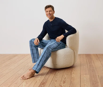 Un homme est assis pieds nus dans un fauteuil, portant un pull bleu marine et un pantalon à carreaux bleus.