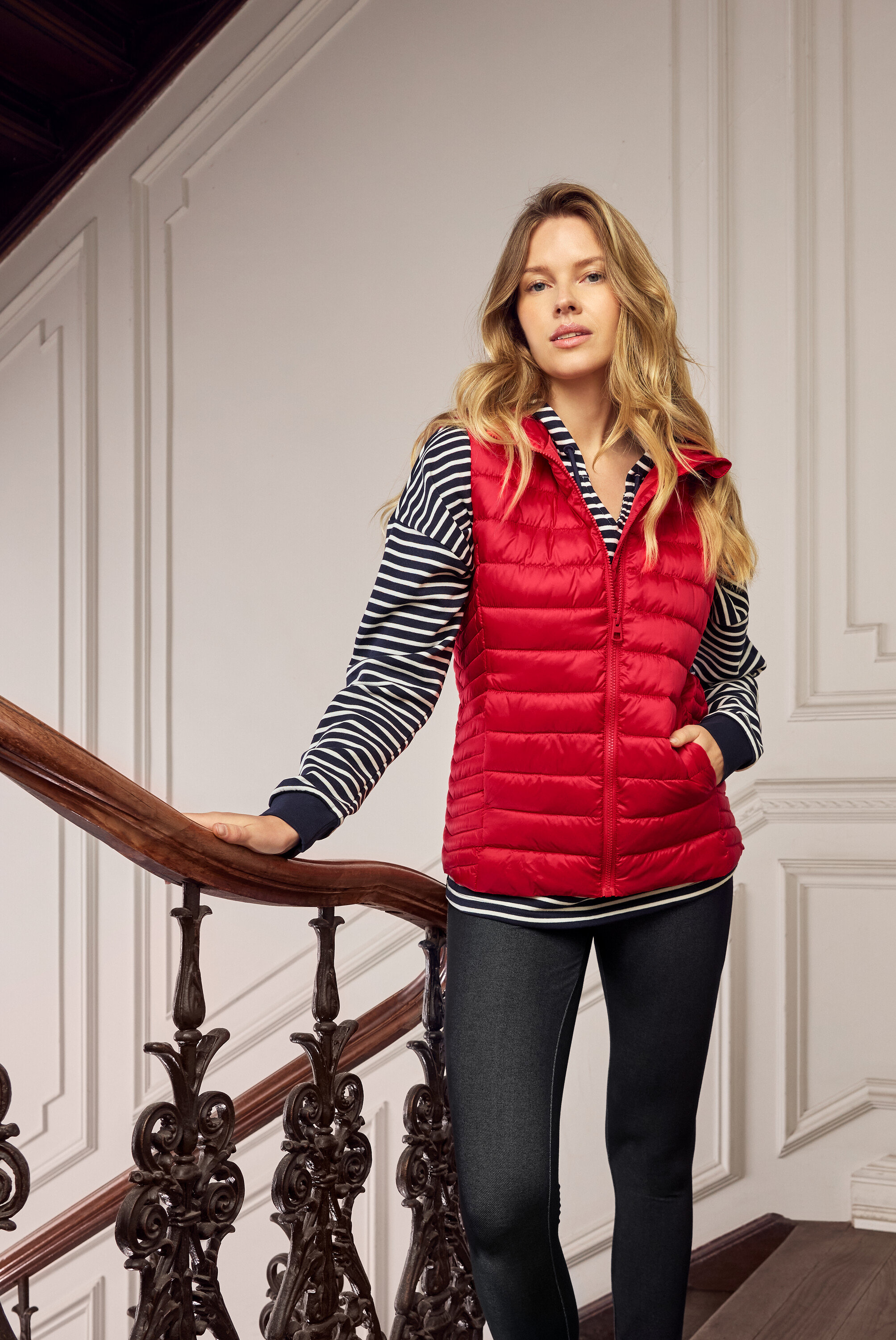 Femme blonde portant un gilet matelassé rouge, un haut rayé bleu et blanc et un jean foncé dans un escalier.
