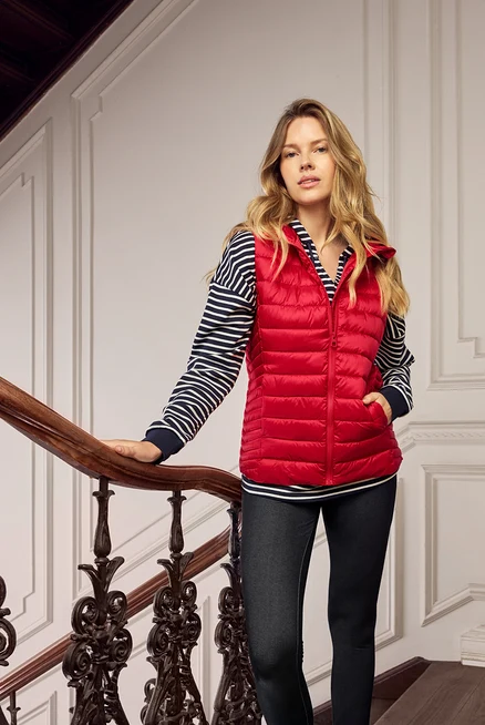 Femme blonde portant un gilet matelassé rouge, un haut rayé bleu et blanc et un jean foncé dans un escalier.