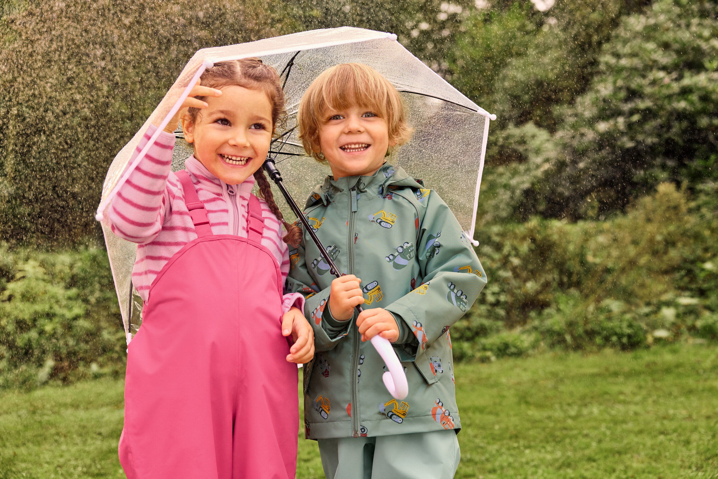 Deux enfants souriants, une fille en salopette rose et un garçon en veste verte avec motif d'excavatrice, se tiennent sous un parapluie transparent sous la pluie.