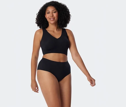 Une femme souriante aux cheveux bouclés foncés porte un haut de soutien-gorge noir et un slip maxi invisible noir SCHIESSER sur fond blanc.