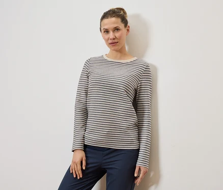 Une femme portant un chignon porte un haut à manches longues rayé gris et blanc et un pantalon bleu marine devant un mur blanc.