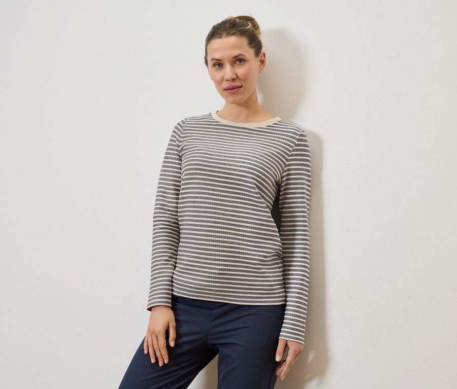 Une femme portant un chignon porte un haut à manches longues rayé gris et blanc et un pantalon bleu marine devant un mur blanc.