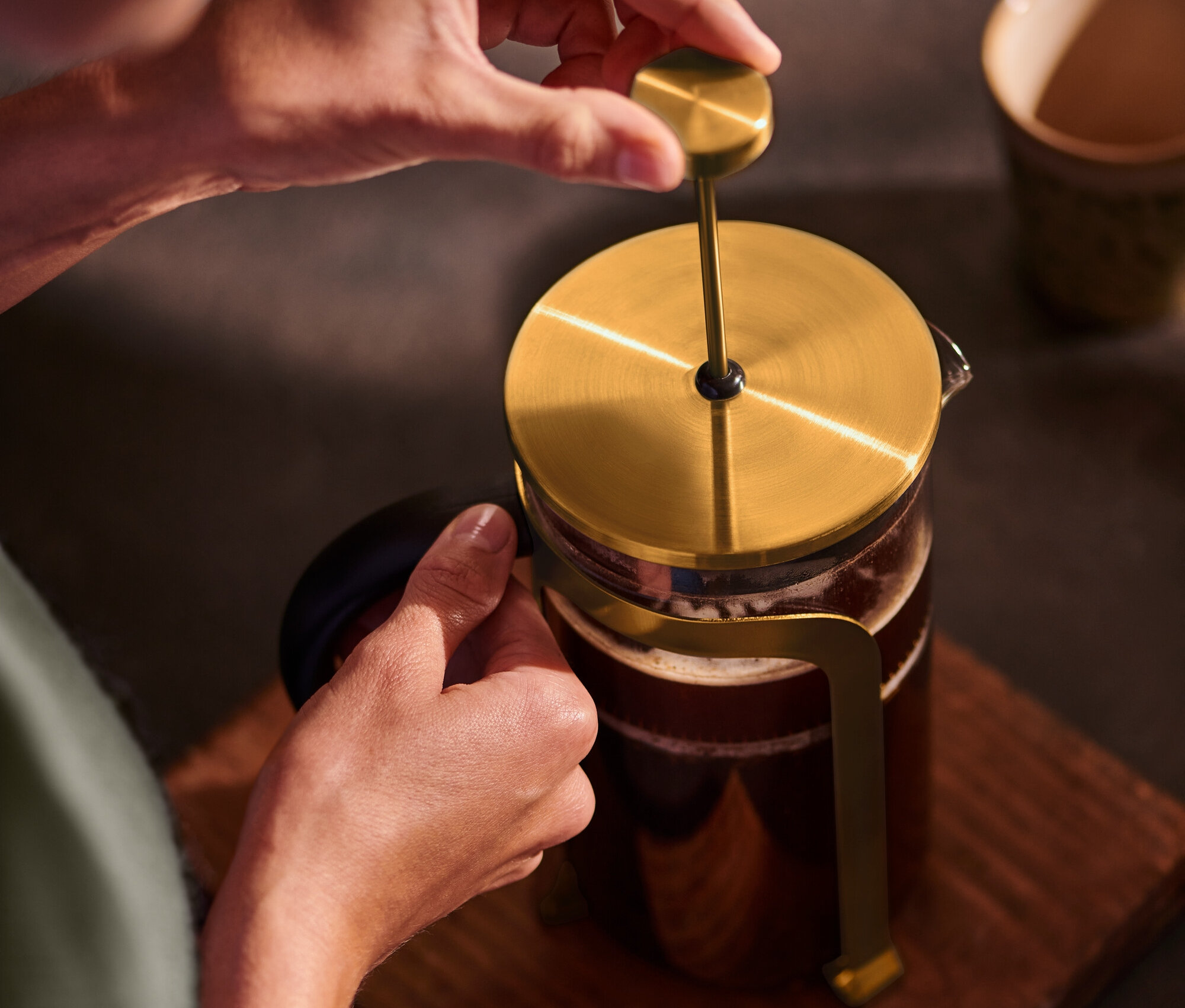 Une personne appuie sur une cafetière à piston dorée de 800 ml pour 6 tasses sur un socle en bois.