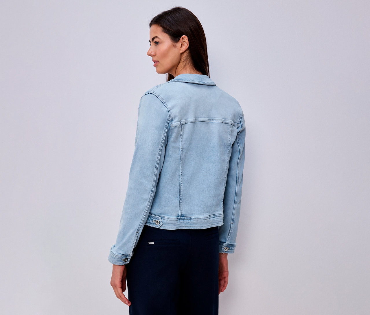 Femme vue de dos, portant une veste en jean bleu clair et une longue jupe bleu marine.