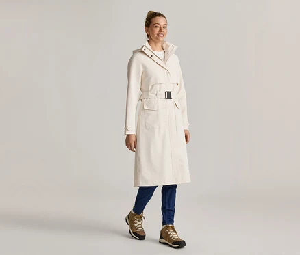 Femme portant un long manteau crème ceinturé, un jean bleu et des bottes marron.