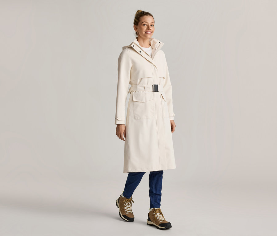 Femme portant un long manteau crème ceinturé, un jean bleu et des bottes marron.