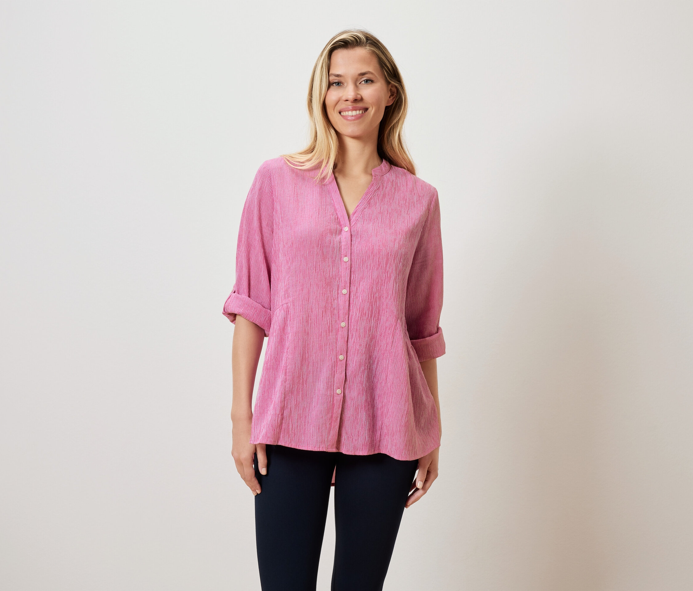 Une femme blonde porte un chemisier rose et un pantalon foncé.