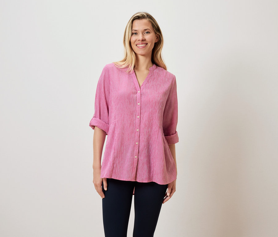 Une femme blonde porte un chemisier rose et un pantalon foncé.