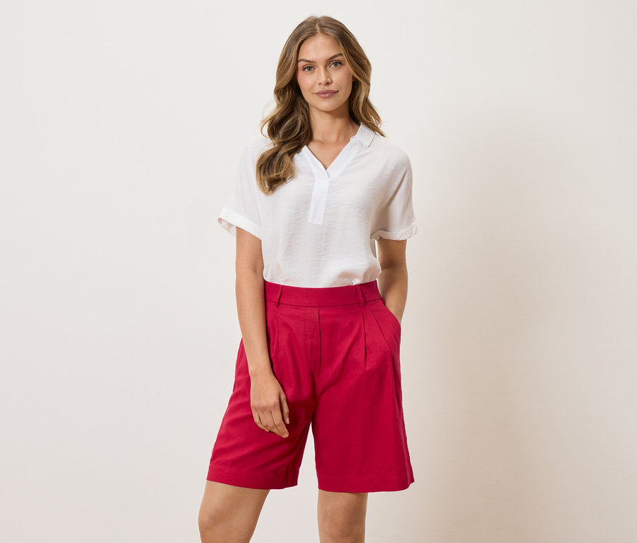 Une femme portant un chemisier blanc et un short rouge se tient devant un fond clair.