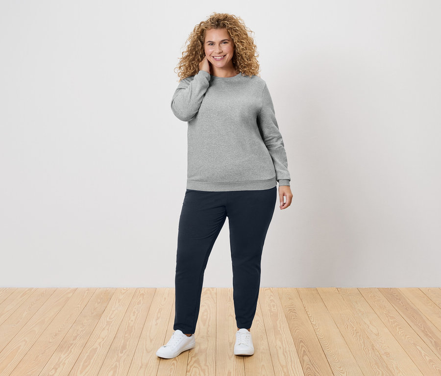 Femme aux cheveux bouclés portant un pull gris et un pantalon bleu marine.
