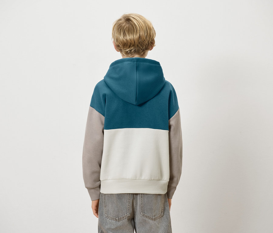 Vue arrière d'un enfant aux cheveux blonds portant un sweat à capuche color block bleu, gris et blanc et un jean gris.