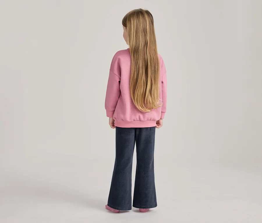 Jeune fille de dos, avec de longs cheveux blonds, portant un sweat-shirt rose et un pantalon évasé bleu foncé.
