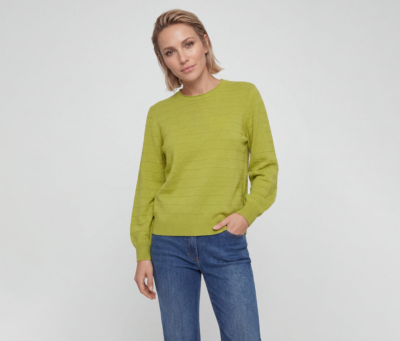 Femme aux cheveux blonds courts portant un pull vert citron à rayures horizontales texturées et un jean bleu, regardant la caméra, une main dans sa poche.