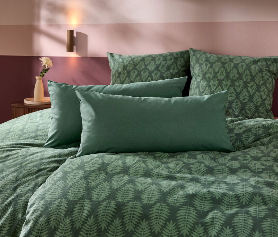 Un lit avec une parure de lit verte à motif de feuilles de fougère et quatre oreillers. En arrière-plan, un mur bicolore dans des tons de violet et de rose, avec une table de chevet, un vase, une fleur et une applique murale.