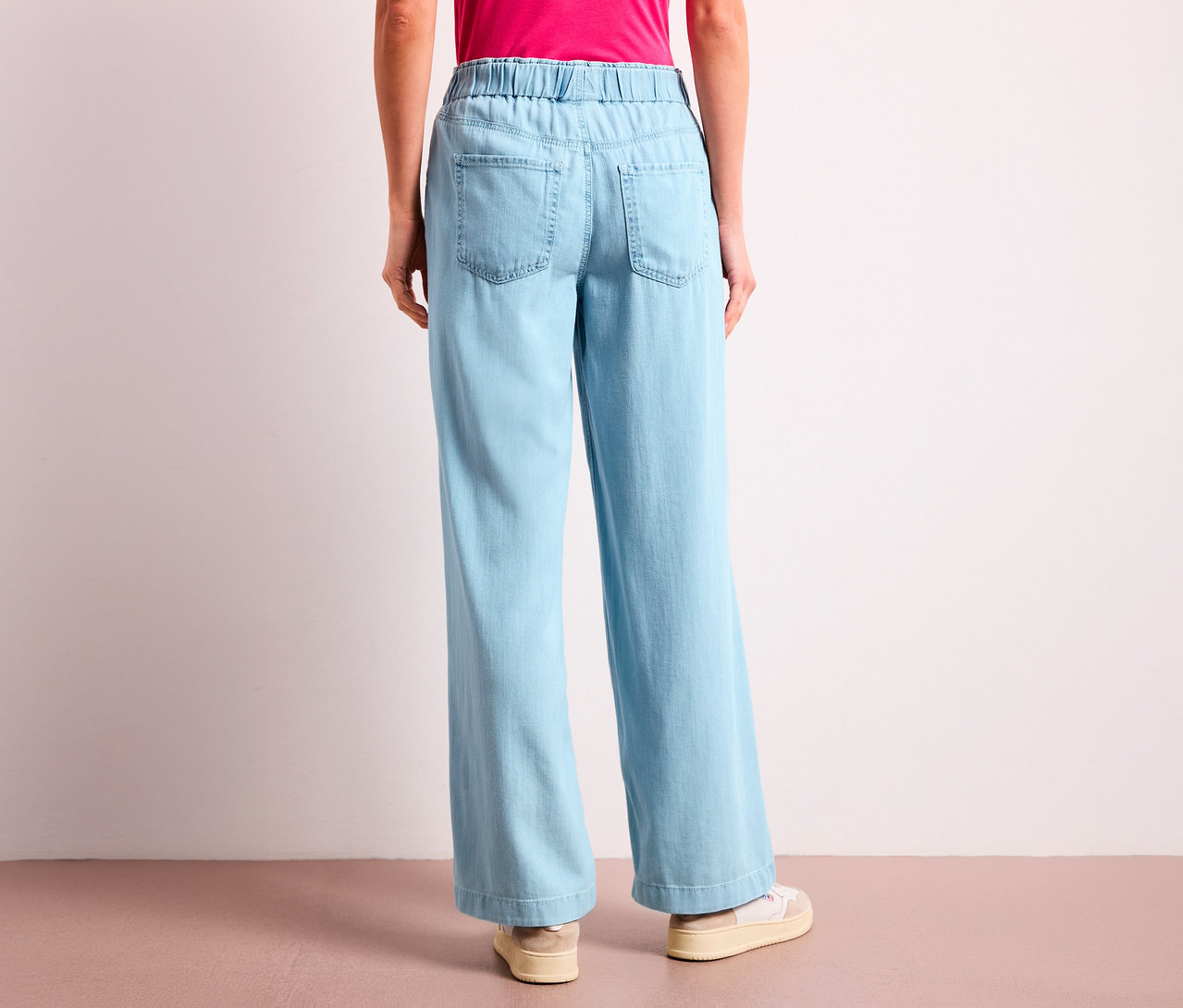 Une personne de dos portant un jean Street One « Emee » bleu clair à jambes larges, avec une taille élastique et des poches arrière. Elle porte aussi un haut rose et des baskets claires.