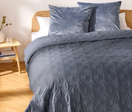 Un lit avec une tête de lit en rotin clair, recouvert d'un couvre-lit matelassé bleu foncé avec un motif géométrique circulaire. Deux oreillers en velours bleu clair reposent contre la tête de lit. À gauche, une table de chevet en bois clair supporte un vase blanc avec une plante et un verre d'eau. La pièce a un parquet en bois clair.