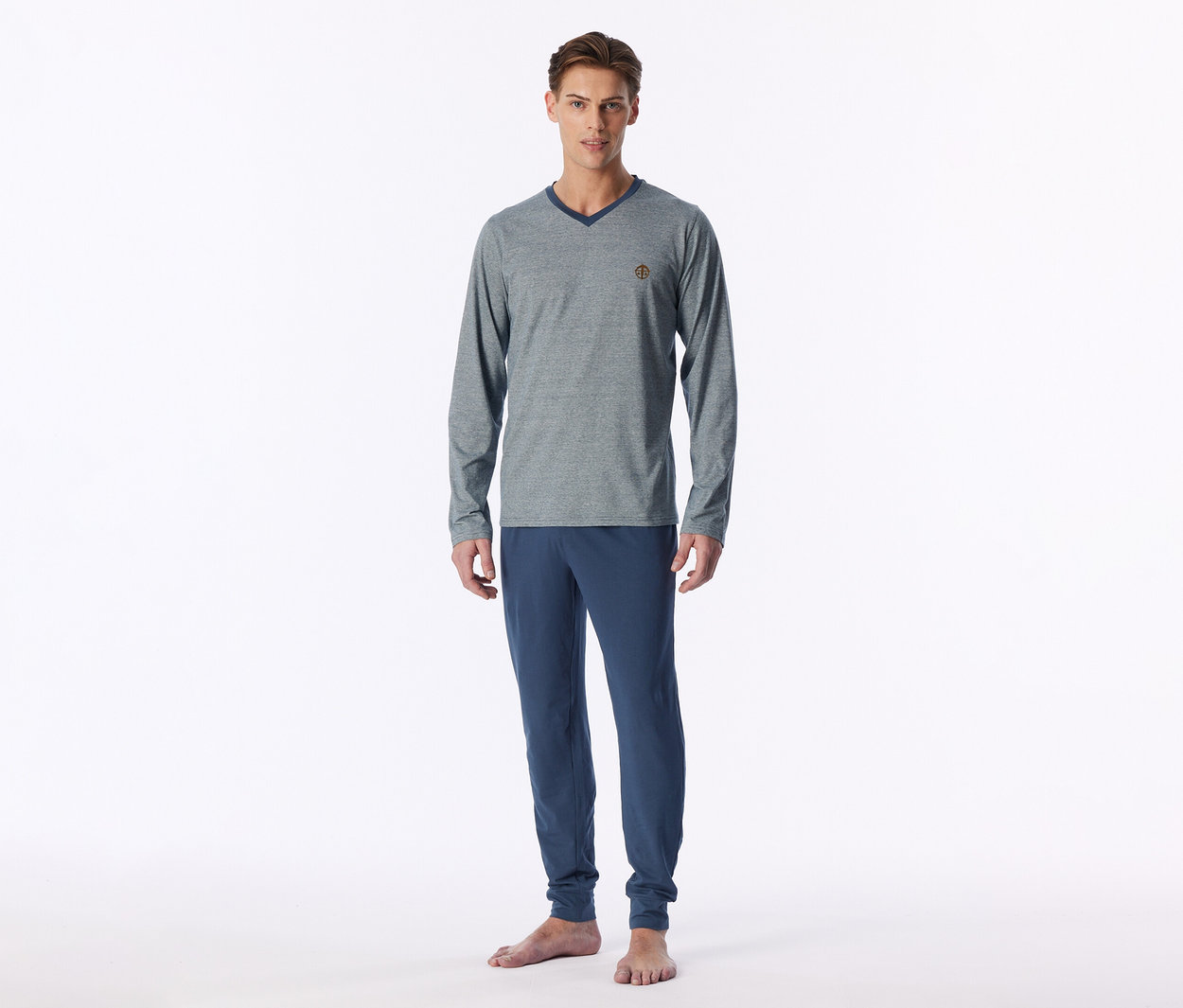 Un homme se tient pieds nus, portant un t-shirt gris à manches longues et col en V avec un col bleu foncé et un pantalon de jogging bleu.