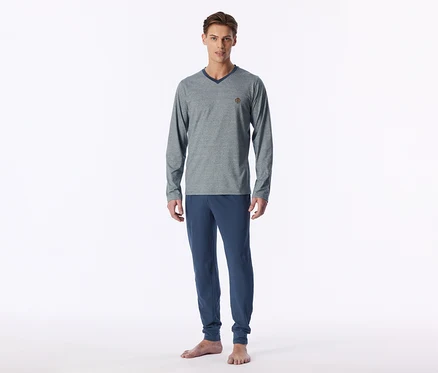 Un homme se tient pieds nus, portant un t-shirt gris à manches longues et col en V avec un col bleu foncé et un pantalon de jogging bleu.