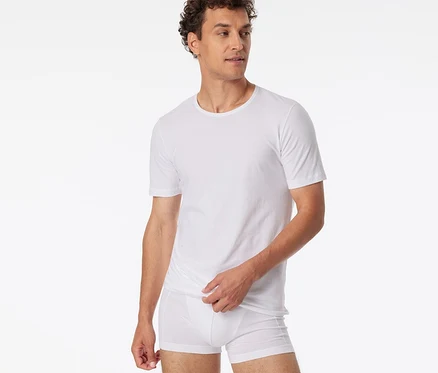 Homme aux cheveux bouclés portant un t-shirt SCHIESSER blanc et un boxer blanc, regardant à droite et remontant l'ourlet de son t-shirt.