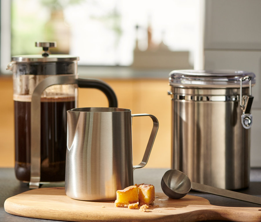 Un verre de café est posé à côté d'une presse française et d'un petit pichet à lait en acier inoxydable sur une planche à découper en bois.