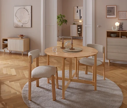 Un coin repas avec une table ronde en bois et trois chaises rembourrées de couleur claire sur un tapis gris. La table est dressée avec des bougeoirs et un verre. À l'arrière-plan, un buffet en bois et une commode sont adossés à des murs neutres ornés d'art abstrait, le tout sur un parquet.
