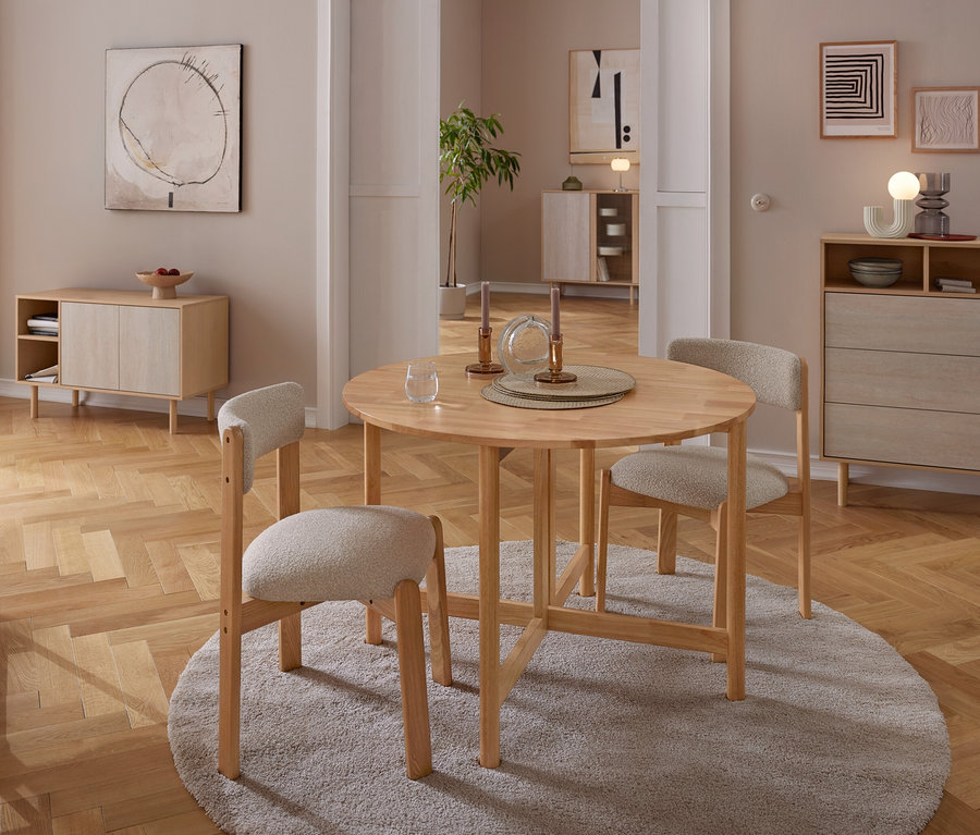 Un coin repas avec une table ronde en bois et trois chaises rembourrées de couleur claire sur un tapis gris. La table est dressée avec des bougeoirs et un verre. À l'arrière-plan, un buffet en bois et une commode sont adossés à des murs neutres ornés d'art abstrait, le tout sur un parquet.