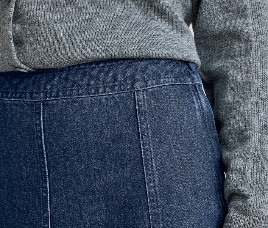 Gros plan d'une personne portant un pull en tricot gris et une jupe en jean bleu foncé avec des coutures visibles.