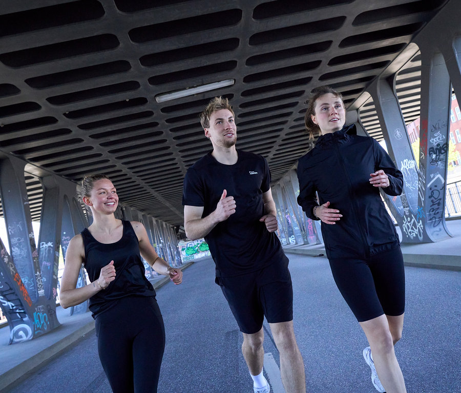 Trois personnes courent sous un pont : Une femme en top de sport noir et leggings de sport « ActivePower » noirs, un homme en T-shirt de sport noir et short de sport noir, et une femme en veste coupe-vent « packable » noire et collant de sport « ActivePower » noir.