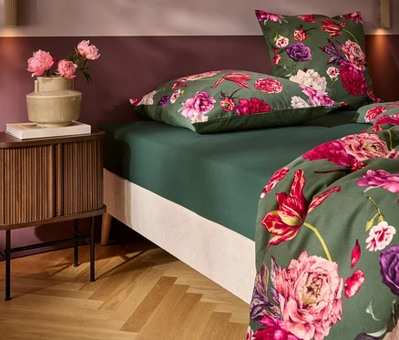 Un lit avec une parure de lit vert foncé et un motif floral vif en rose, fuchsia et violet. À gauche, une table de chevet en bois avec un vase de pivoines roses et un livre. Le mur est bicolore, prune et rose clair, au-dessus d'un parquet en chevrons.